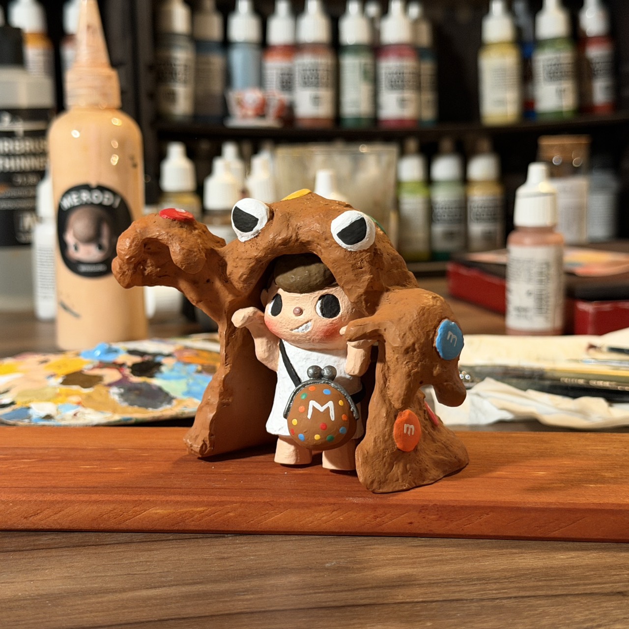 A hand-painted Merodi Cookie Monster figure resting on a wooden strip among studio paint bottles